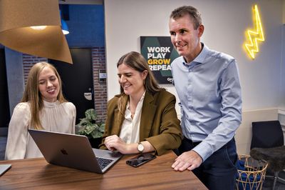 Anne, Katrine, og Stein Erik smiler ved en laptop i et kontor med et neonlys-lyn på væggen.